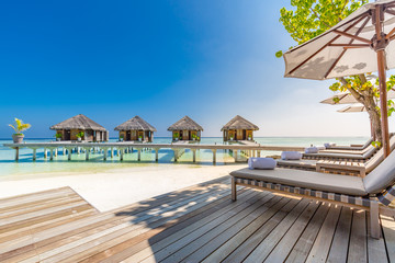 Sunny day on Maldives island, luxury water villas resort jetty and loungers under umbrella. Beautiful sky and clouds and beach background for summer vacation holiday and travel concept