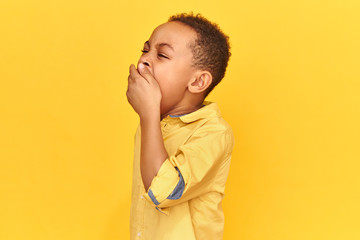 Horizontal shot of exhausted sleepy African schoolboy wearing yellow shirt covering mouth with hand yawning being tired after long wearisome day. Boredom, sleep, bedtime and bedding concept - obrazy, fototapety, plakaty