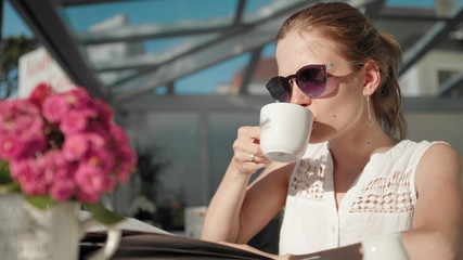 Slender Young Adult Woman with Ponytail and Sunglasses is Drinking Coffee in the Terrace of a Restaurant and Reading Menu in Summer