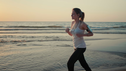 Beautiful Woman is Running along Sea Coast at Sunset and Jogging on Beach with Headphones