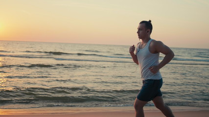 Sporty Hadsome Man is Running along Sea Coast at Sunset while Jogging on Beach with Headphones