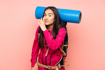 Young mountaineer Indian girl with a big backpack isolated on pink background whispering something