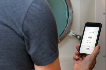 Male cruise passenger looking the world statistics of the coronavirus covid-19 outbreak on a mobile phone over a ship porthole.