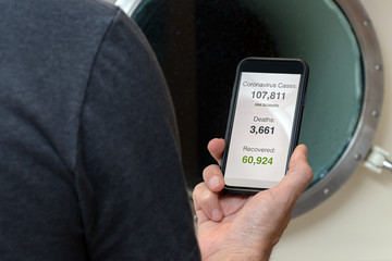 Male cruise passenger looking the world statistics of the coronavirus covid-19 outbreak on a mobile phone over a ship porthole.