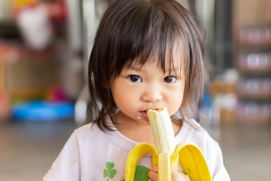 Portrait image of baby 1-2 years old. Happy Asian child girl enjoy eating and biting a banana with sweet smiling. Food and healthy kids concept.