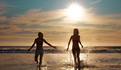 Silhouette of two beautiful women running on a beach shore with waves in the background at sunset or sunrise.