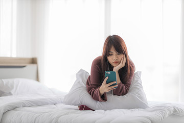 Asian happy woman sitting on bed is enjoying listening to music and watching entertainment from her smartphone with earphones.