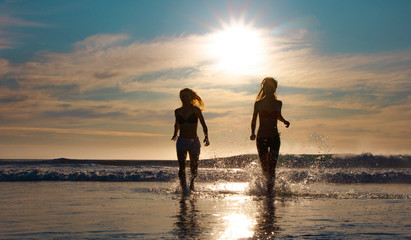 Silhouette of two beautiful women running on a beach shore with waves in the background at sunset or sunrise.