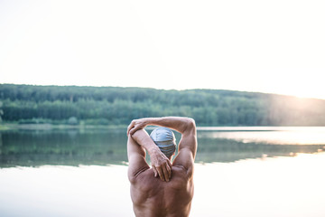 Rear view of senior man standing by lake outdoors before swimming, stretching.