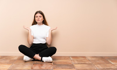 Ukrainian teenager girl sitting on the floor having doubts while raising hands