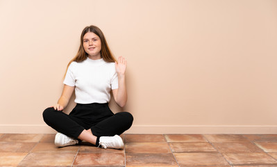 Ukrainian teenager girl sitting on the floor saluting with hand with happy expression