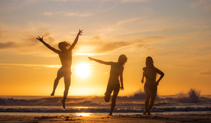 Silhouette of three beautiful women having fun running and jumping on a beach at sunset or sunrise.