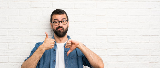 Handsome man with beard over white brick wall making good-bad sign. Undecided between yes or not