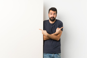 Young handsome man with beard holding a big empty placard pointing to the laterals having doubts