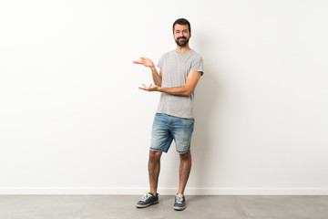A full-length shot of handsome man with beard extending hands to the side for inviting to come - obrazy, fototapety, plakaty