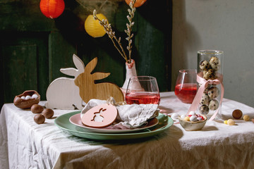 Easter table setting with wooden rabbit decoration, chocolate eggs and sweets, glasses of pink cocktail drinks, empty plates on white tablecloth. Lighting garlands. Holiday dinner.
