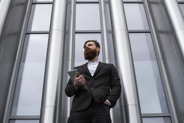 Modern Young bearded Business man standing with a digital tablet. Young hipster businessman holding tablet in hands outdoor. Working online with a tablet while standing outside on an office building.