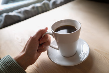 Black coffee in a white mug and saucer being held by a woman. The cuff of a cord jacket can be seen. the Table is wooden and the scene is lit with natural window light