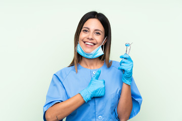 Woman dentist holding tools over isolated green background giving a thumbs up gesture