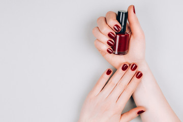 Beautiful dark red manicure with a bottle of nail polish in hands on a grey background. Procedures...
