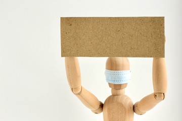 Cardboard sign with space for text in the hands of a wooden doll or mannequin in a medical mask. Concept of anti racism, anti xenophobia, the social situation around the Coronavirus or Covid-19