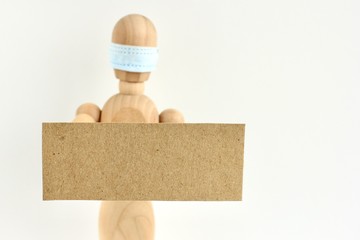 Cardboard sign with space for text in the hands of a wooden doll or mannequin in a medical mask. Concept of anti racism, anti xenophobia, the social situation around the Coronavirus or Covid-19