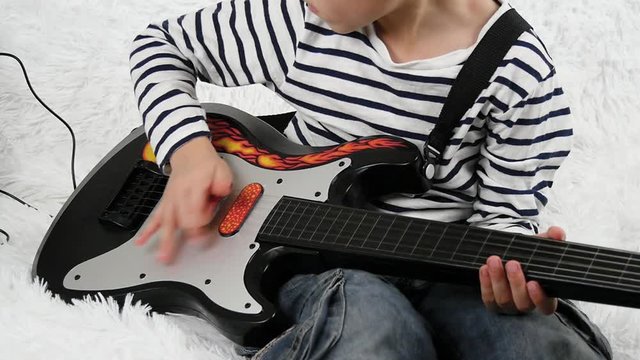 A preschool child spends time playing a small electric guitar for children. The child touches the strings of the guitar with his fingers , imagining the musician. Happy child.