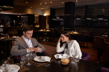offended caucasian girl and young guy sit in restaurant, they don't talk with each other. beautiful couple came to celebrate anniversary, but they had quarrel. man look at smartphone
