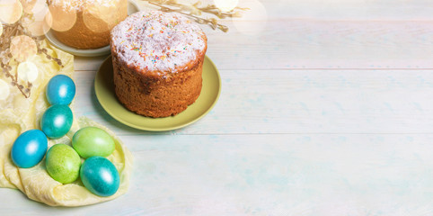Easter festive banner with Easter cakes, colored Easter eggs on yellow cloth, willow twigs, bokeh on painted wooden background. Holiday background. Copy space for text.