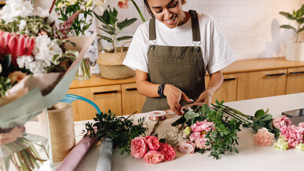 Smiling florist preparing a flower bouquet at her shop. Woman in apron working at counter. - obrazy, fototapety, plakaty