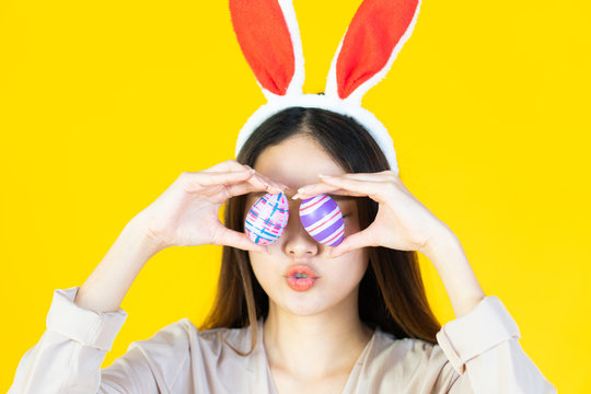 A beautiful young asian woman with smiling face wearing bunny ear, holding multicolored set of easter eggs on the yellow baclground