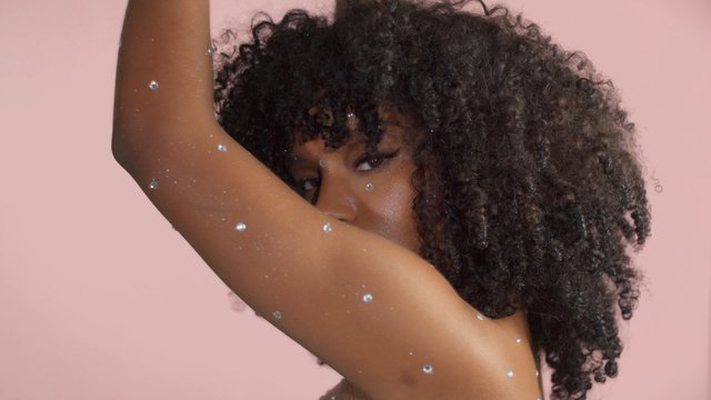 Mixed race black woman with curly hair covered by crystal makeup on pink background in studo dancing lifted her hand to cover her face