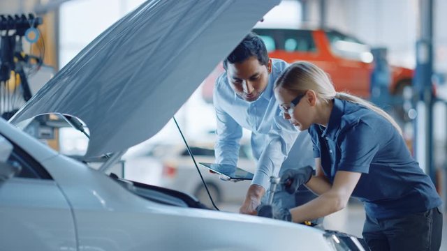 Manager Checks Diagnostics Results on a Tablet Computer and Explains an Engine Breakdown to an Female Mechanic. Car Service Employee Uses a Ratchet to Fix the Engine Component. Modern Clean Workshop.