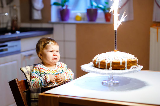 Cute Crying Little Baby Girl Celebrating First Birthday. Child Scared And Afraid Of Firework On Baked Cake, Indoor. Toddler Chil Not Happy