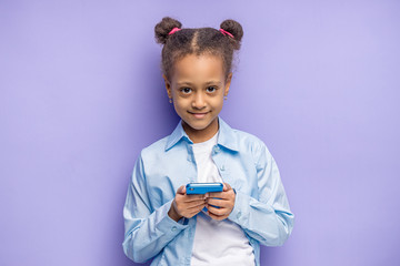 portrait of cute smiling or african kid girl with smartphone isolated over purple background. happy child chatting with friend, she has bunched curly hair, look at camera