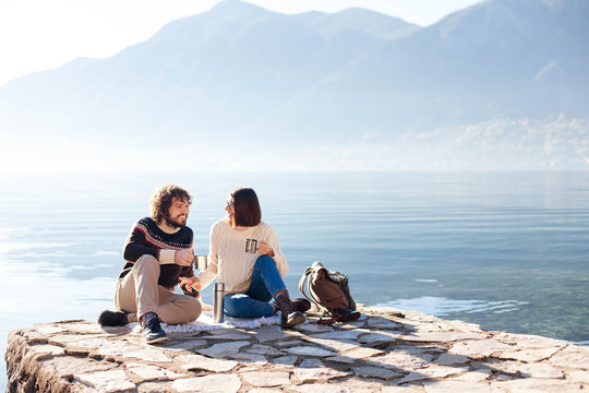 Picnic at winter sea beach. Couple in love drink coffee. Young travelers sitting outdoors. Happy man and woman enjoying nature, relaxation, togetherness and traveling. Romantic lifestyle moment.