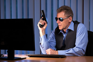 Criminal businessman sitting with pistol in his hand at a desk with monitor in a dark office by...