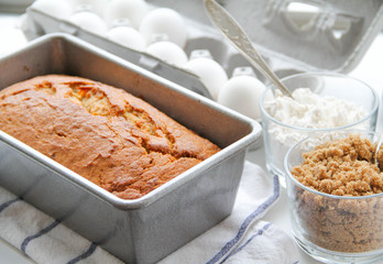 Banana bread in a metal loaf pan is on a kitchen towel. There are eggs, and brown sugar and flour in small glasses near the pan. Back view. 