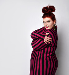 Plus size redhead model in striped dress, earrings. She is hugging herself while posing sideways isolated on white background
