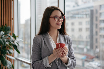 Portrait of happy successful woman in formal clothes and stylish eyeglasses planning start up. Young confident businesswoman standing in modern office near window, holding cup of coffee and smiling