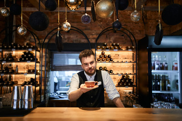 Close up of handsome bearded barista holding cup of coffee in hand looking at the camera at the cafe, close up photo.job, profession, occupation