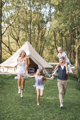 Fototapeta premium Happy Caucasian family, father mother and two little daughters, having fun outdoors in park or camping, enjoying time and laughing together, holding hands and running. Love and parenthood concept