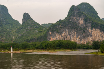 Guilin, China - May 10, 2010: Along Li River. Long shot of white and red navigation beacon standing at edge of water with forest covered karst mountains and brown cliff in back under rain cloudscape.