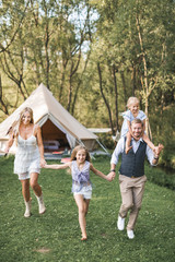 Fototapeta premium Young family in boho casual wear, dad, mom and two daughters holding hands and running and jumping in a green park, big wigwam tent on the background. Family walk, outdoors