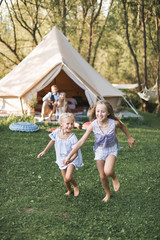 Fototapeta premium Beautiful young family spending time together in summer park or meadow, having a picnic. Young parents sitting near wigwam, while two little girls daughters running and laughing