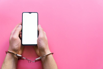 Men's hands in handcuffs hold a smartphone with blank screen on a pink background. The concept of internet and gadget dependency.