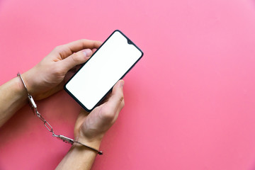 Men's hands in handcuffs hold modern smartphone on a pink background. The concept of internet and gadget dependency.