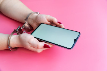 Woman hands in handcuffs hold a modern smartphone on a pink background. The concept of internet and gadget dependency.