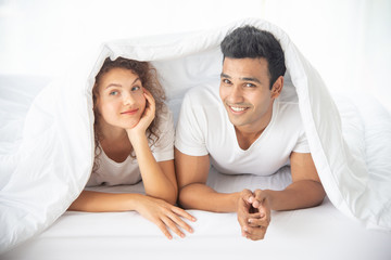 Funny couple lying, talking, hiding in white blanket and looking at the camera  on white bed  in the bedroom. Relation in family. Relaxing in the morning.