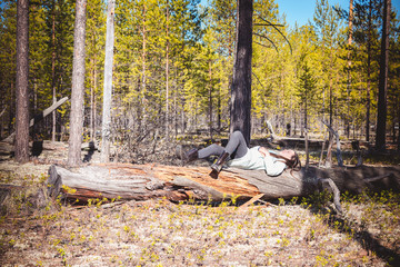 Tired girl in light jacket after hike sleeps with her eyes closed on fallen tree in pine forest.